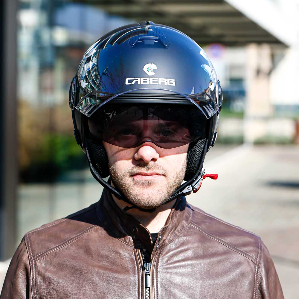 A man wearing the matte black Caberg Riviera V4X Mia helmet, seen from the front with the clear visor lifted, revealing the integrated sun visor and floral graphics.